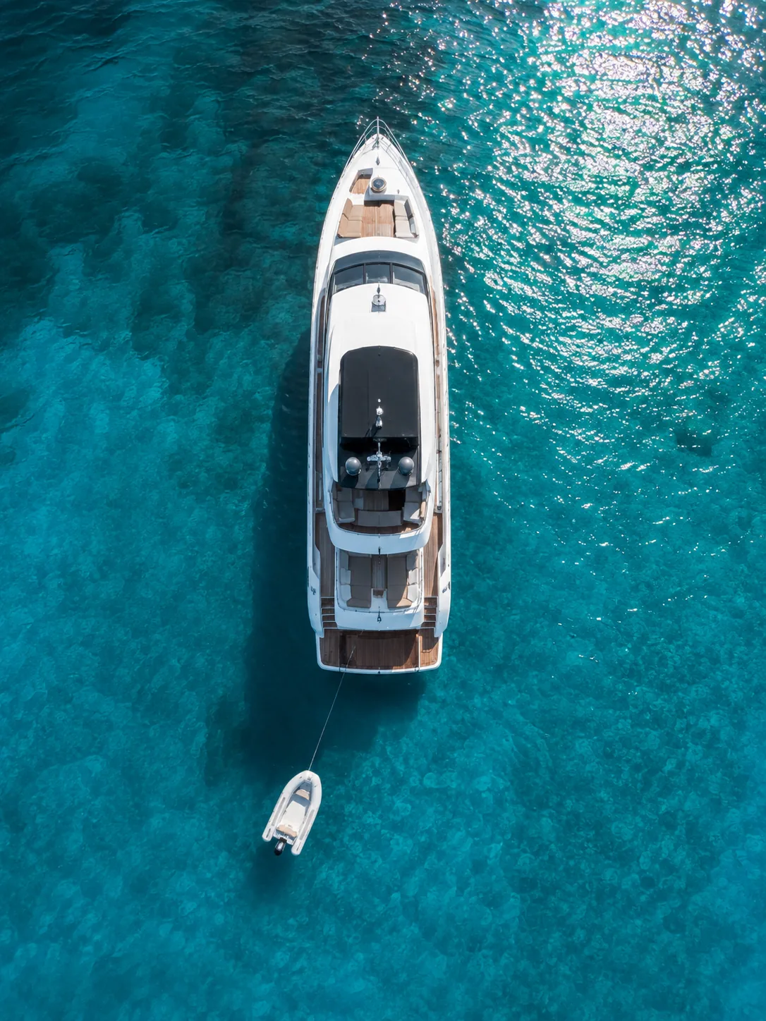 Aerial view of a motor yacht at anchor in clear turquoise water, its tender astern